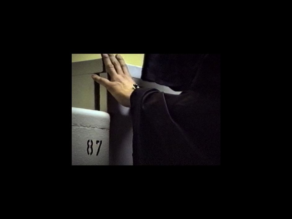 Archival material: Black-framed photograph of an arm wrapped in black fabric reaching for a white metal cabinet. Next to it, another metal object is recognisable, on which the number 87 is written.