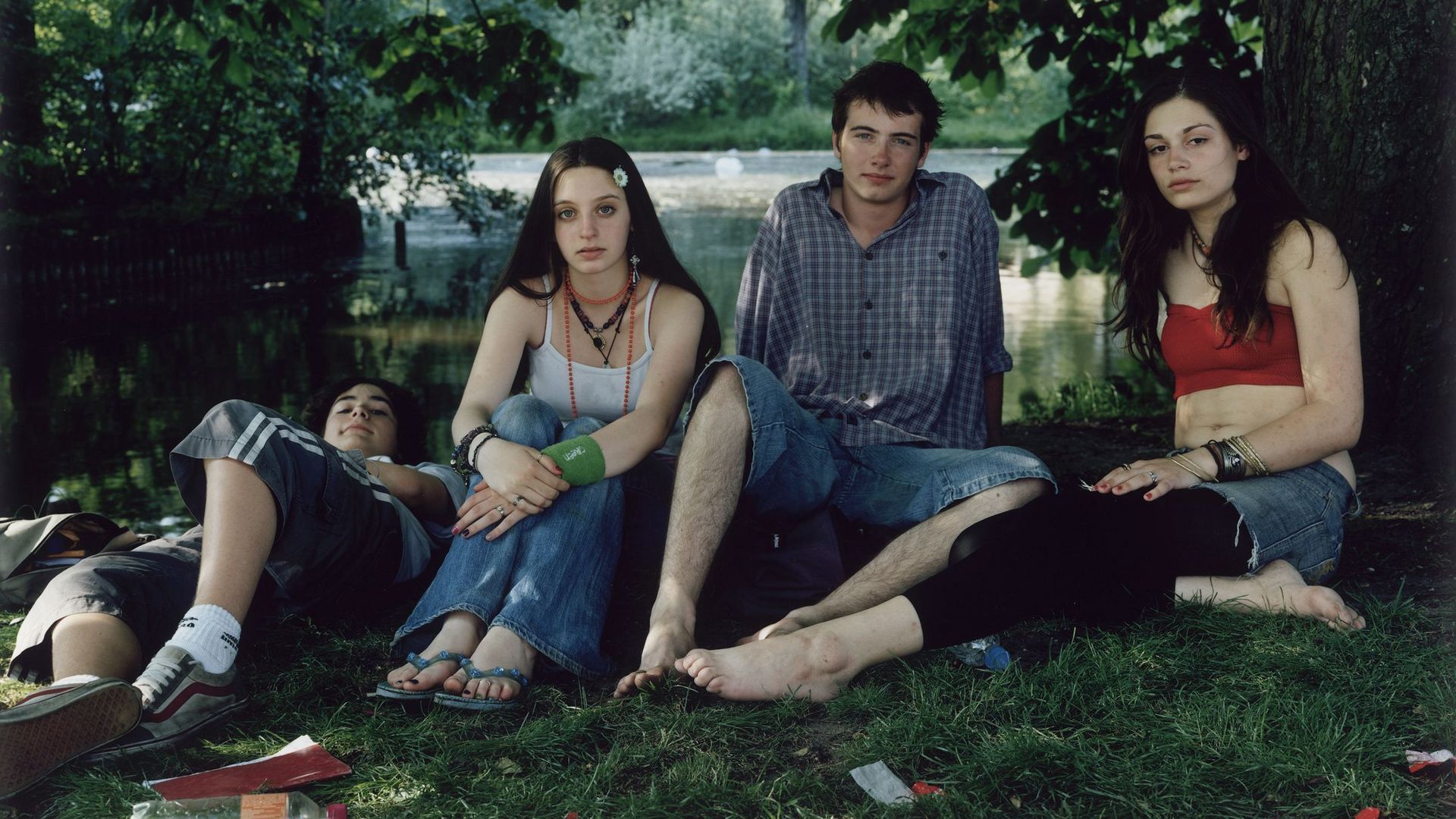 Fotografie: Vier junge Personen sitzen bzw. liegen in Sommerkleidung in einem Park. Ihr direkt ist direkt in die Kamera gerichtet.