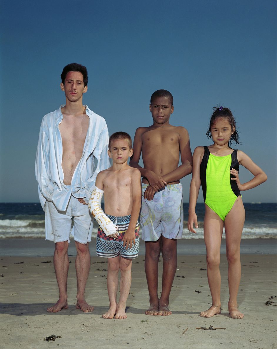 Fotografie: Vier junge Personen stehen in Badekleidung am Strand und blicken direkt in die Kamera.