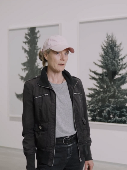 Videostill: Eine Person mit rosa Kappe und schwarzer Jacke steht vor vier eingerahmten Fotografien von Nadelbäumen.