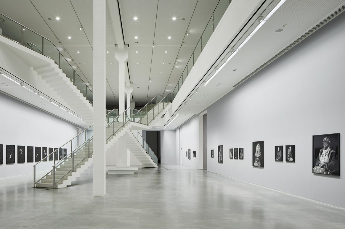 Foto: Zweistöckiger Ausstellungsraum mit Kunstwerken an den Wänden, in der Mitte zwei sich kreuzende Treppenaufgänge.