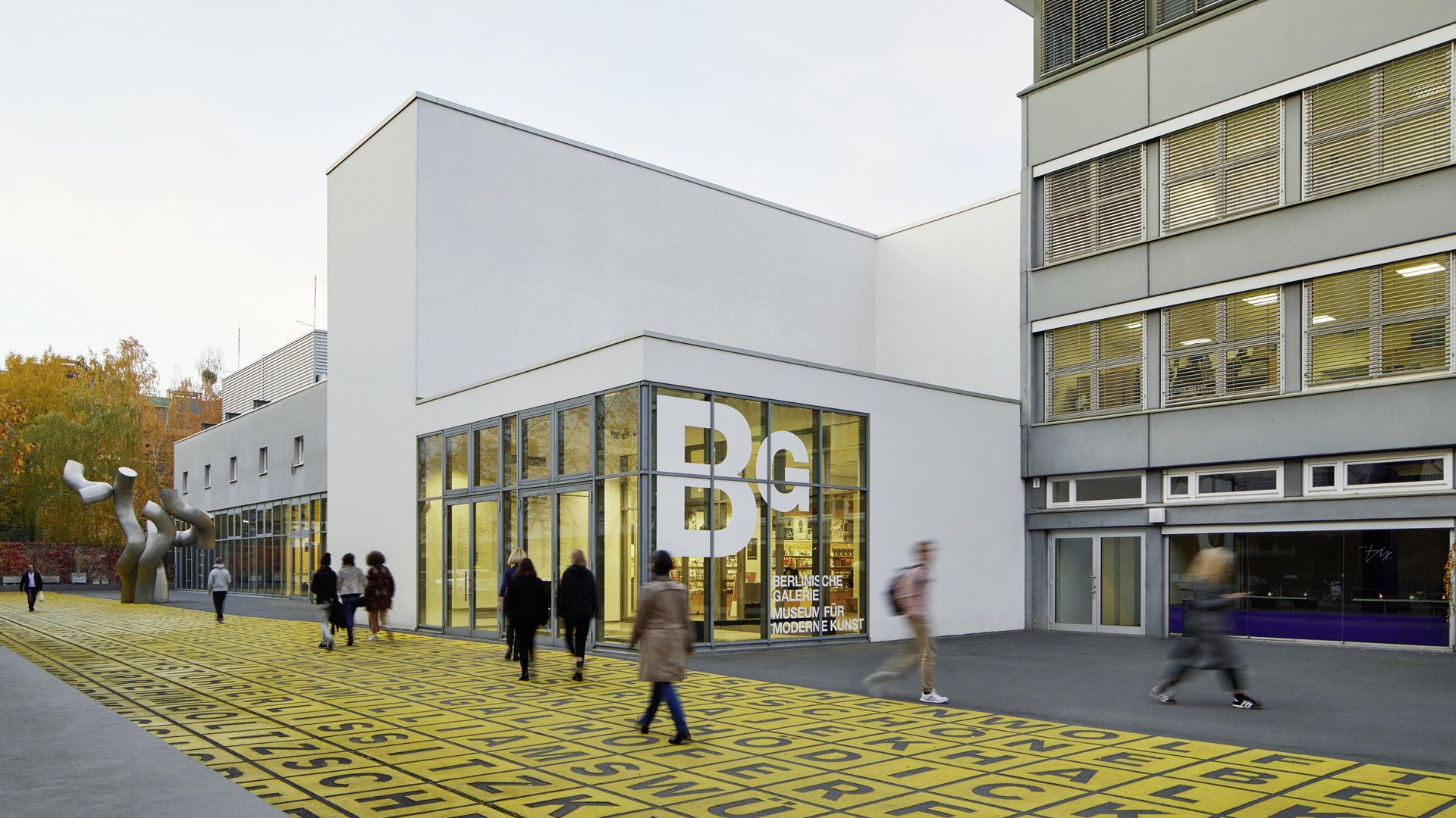 Foto: Gebäude mit Flachdach und Glasfront, auf der das Logo der Berlinischen Galerie abgebildet ist. Davor Menschen auf einem Platz mit Metallskulptur und Bodenkunstwerk.