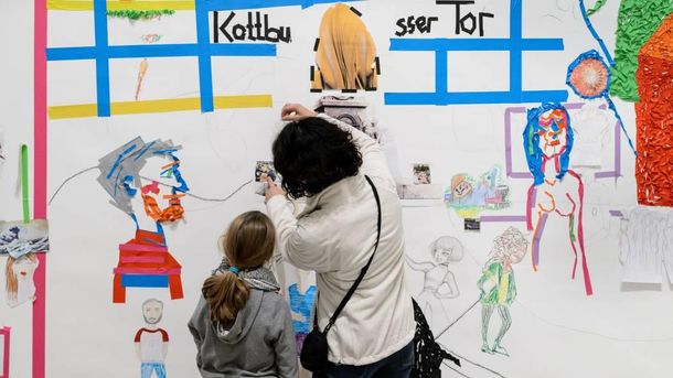 Foto: Ein Kind und eine Erwachsene Person stehen vor einen großen Wandkunstwerk von Kindern.