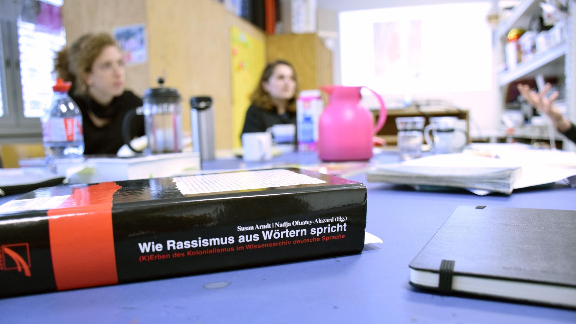 Foto: An einem Tisch sitzen zwei Menschen an einem Tisch mit Arbeitsmaterialien. Im Vordergrund ein Buch mit dem Titel "Wie Rassismus aus Wörtern spricht"