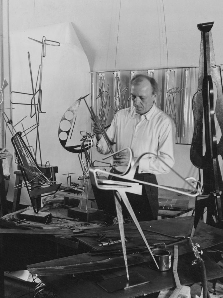 Black and white photograph of Hans Uhlmann in his studio. The artist is surrounded by his metal sculptures in progress.