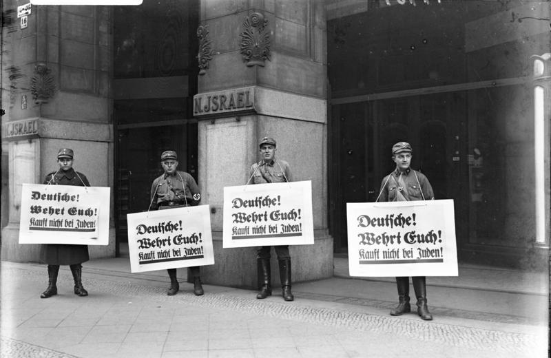 Fotografie: Männer in Uniform vor einem Gebäude, sie halten Schilder hoch. Aufschrift: „Deutsche! Wehrt euch! Kauft nicht bei Juden"
