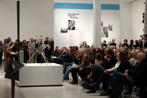 Photo: Side view of the staircase hall of the Berlinische Galerie, where many people sit in rows of chairs and watch the four speakers, Thomas Köhler, Ilka, Voermann, Ursula Müller and Christine van Haaren. 