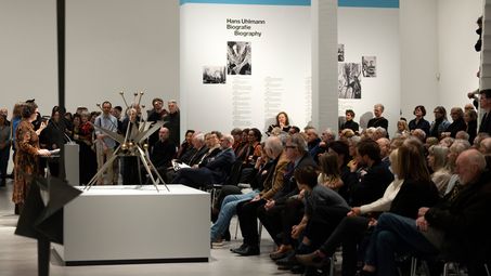 Foto: Seitlicher Blick in die Treppenhalle der Berlinischen Galerie, wo viele Personen in Stuhlreihen sitzen und die vier Redner*innen, Thomas Köhler, Ilka, Voermann, Ursula Müller und Christine van Haaren, ansehen. 