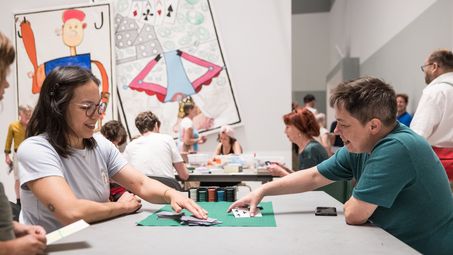 Foto: Einige Personen sitzen an Tischen im Projektraum der Berlinischen Galerie, spielen Kartenspiele, basteln und unterhalten sich.