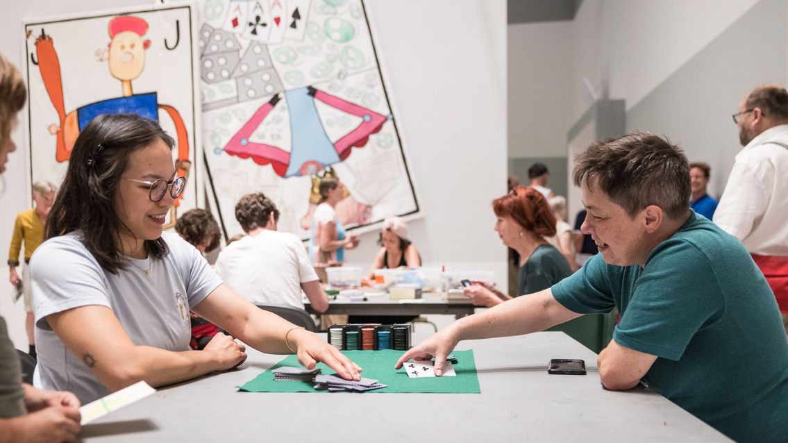 Photo: Some people are sitting at tables in the project room of the Berlinische Galerie, playing card games, doing handicrafts and chatting.