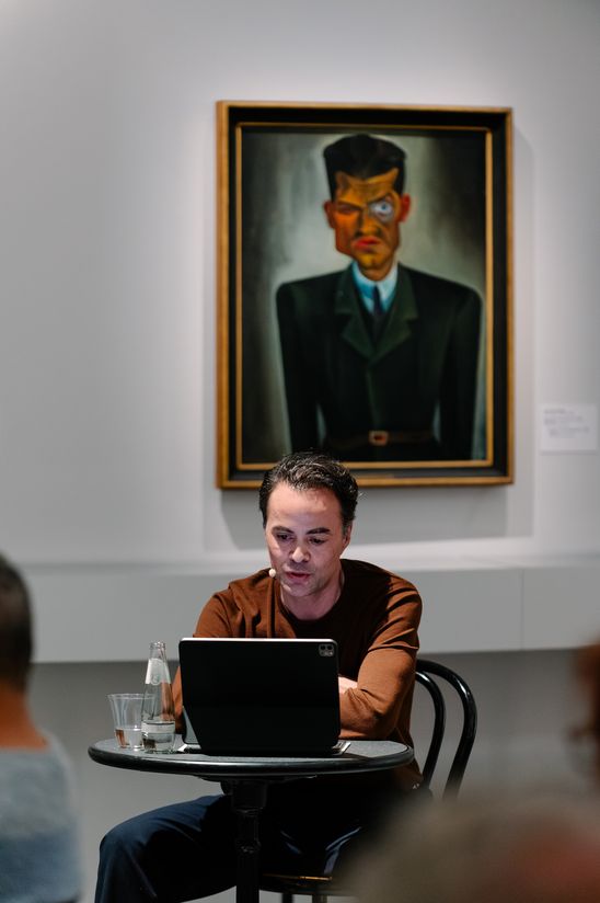 Photo: Nikolai Kinski sits at a small table in front of a painting depicting Raoul Hausmann.