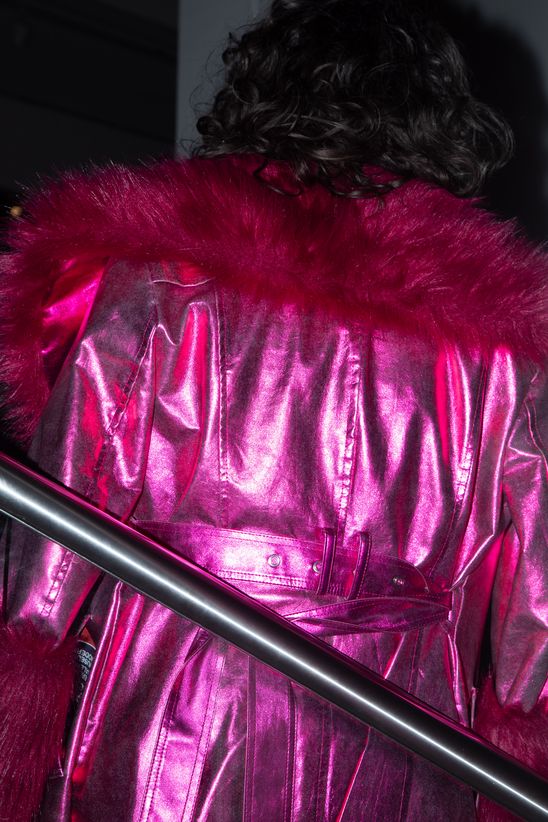 Foto: Rückenansicht einer Person, die in einem leuchtend metallic-pinkfarbenen Mantel am Treppengeländer lehnt.