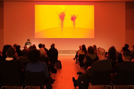 Photo: Monira Al Qadiri stands at a lectern in front of an audience. The room is bathed in warm, orange light from her presentation projected onto the wall.