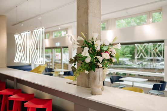 Photo: View of the interior of the museum café at the Berlinische Galerie. The café's logo hangs in illuminated letters above a simple counter with bright red bar stools. At the back of the room there are more tables with chairs and a large window front. 