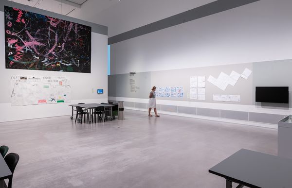 Foto. Ein hoher Raum mit Tischen und Stühlen. An der linken Wand ein großes Kunstwerk aus Papier, an der rechten Wand viele kleine.