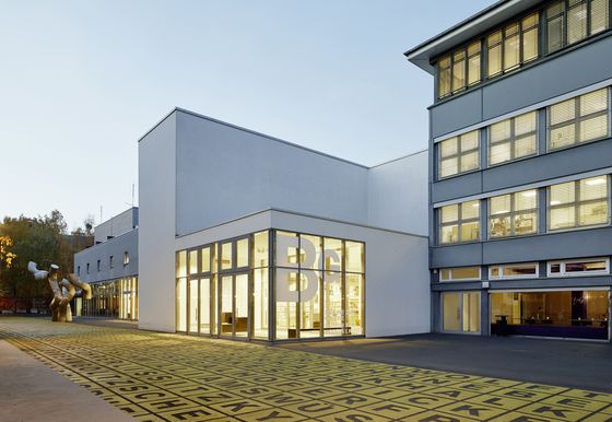 Foto: Gebäude mit Flachdach und Glasfront, auf der das Logo der Berlinischen Galerie abgebildet ist. Davor ein Platz, darauf Menschen, eine Metallskulptur und Bodenkunstwerk.