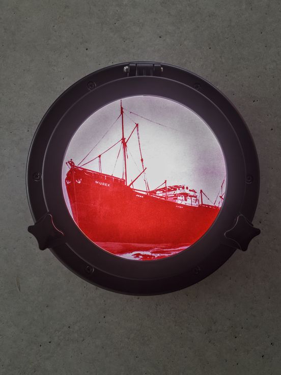 Photo: View of a round window (porthole) in which a large ship can be seen in bright red colour on a white and pink background. ‘Murex’ is written in capital letters on the bow of the ship.