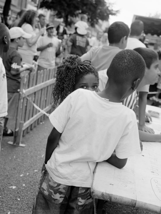 Black and white photograph: In a square format, the photograph shows several people standing mainly with their backs to the camera, while a young, female figure looks directly into the camera.