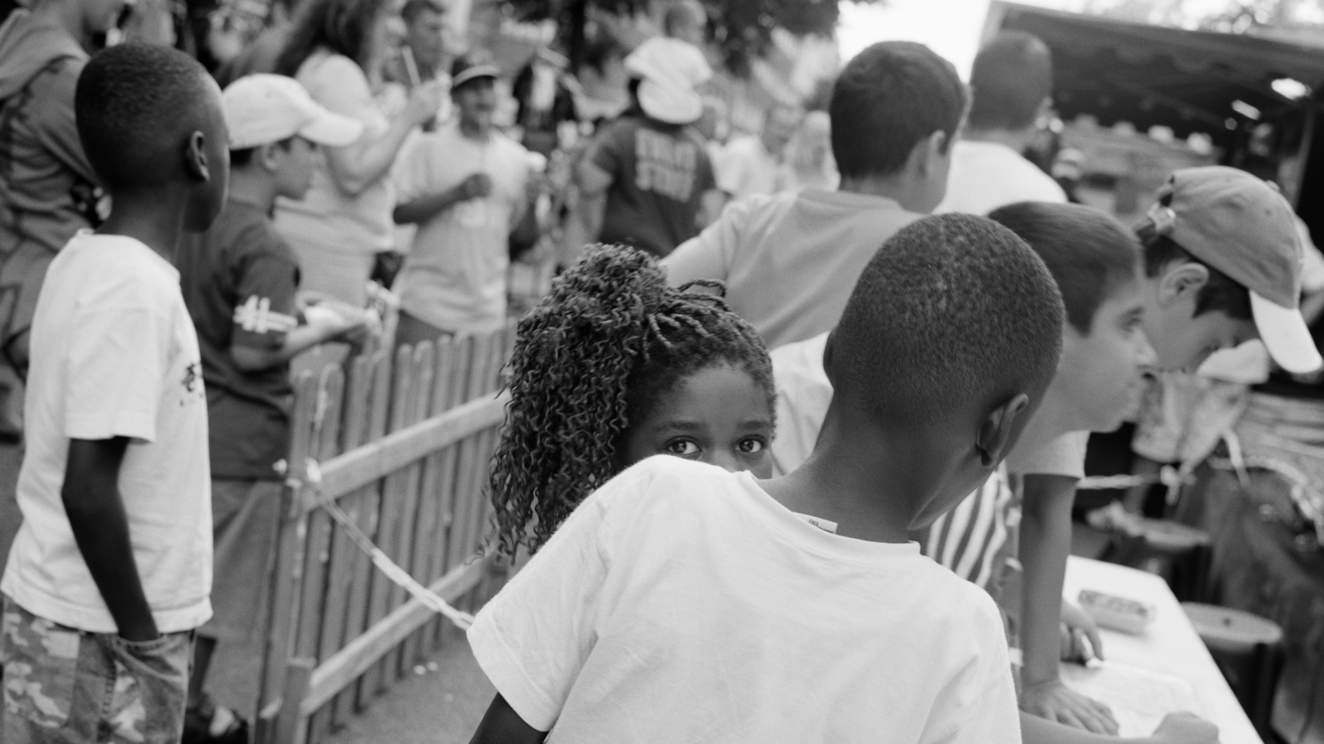 Schwarz-weiß-Fotografie: Im quadratischen Format zeigt die Fotografie mehrere Personen, die vorwiegend mit dem Rücken zur Kamera stehen, während eine junge, weiblich gelesene Person direkt in die Kamera blickt.