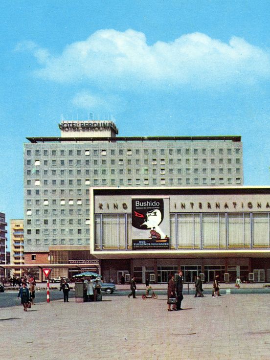 Street scene with several buildings: In the background, a tall rectangular hotel building with many uniform windows and a sign reading “Hotel Berolina” on the roof. In front of it is a wide, low building with large window fronts and a movie poster on the facade. On the right side of the image, a modern building with large glass fronts and signage on the roof is visible. People are walking across the plaza, and some cars are parked or driving in the background. The sky is clear and blue.