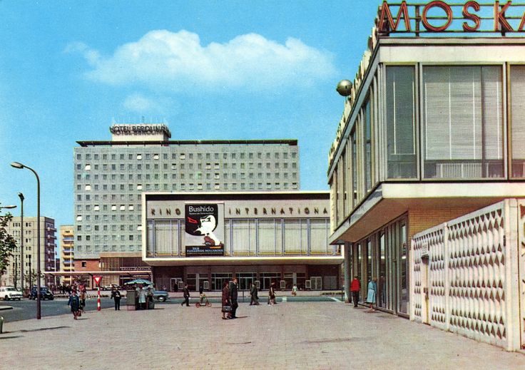 Straßenszene mit mehreren Gebäuden: Im Hintergrund steht ein hohes, rechteckiges Hotelgebäude mit vielen gleichförmigen Fenstern und einem Schild „Hotel Berolina“ auf dem Dach. Davor befindet sich ein breiter, niedriger Bau mit großen Fensterfronten und einer Filmwerbung an der Fassade. Rechts im Bild ist ein modernes Gebäude mit großen Glasfronten und einem Schriftzug auf dem Dach zu sehen. Auf dem Platz davor gehen verschiedene Personen, einige Autos stehen oder fahren im Hintergrund. Der Himmel ist klar und blau.