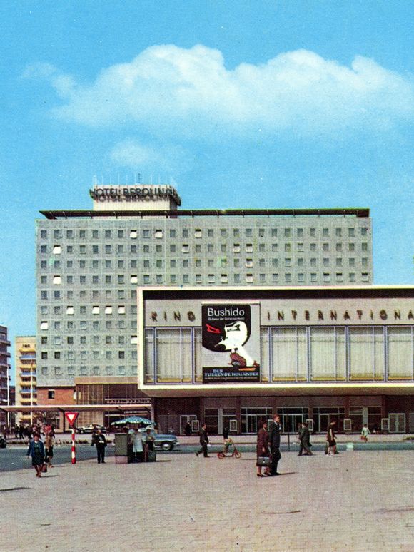 Street scene with several buildings: In the background, a tall rectangular hotel building with many uniform windows and a sign reading “Hotel Berolina” on the roof. In front of it is a wide, low building with large window fronts and a movie poster on the facade. On the right side of the image, a modern building with large glass fronts and signage on the roof is visible. People are walking across the plaza, and some cars are parked or driving in the background. The sky is clear and blue.