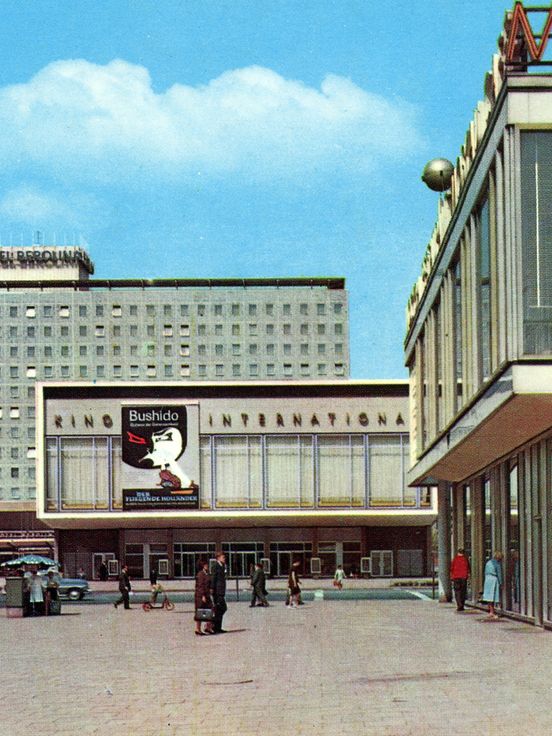 Street scene with several buildings: In the background, a tall rectangular hotel building with many uniform windows and a sign reading “Hotel Berolina” on the roof. In front of it is a wide, low building with large window fronts and a movie poster on the facade. On the right side of the image, a modern building with large glass fronts and signage on the roof is visible. People are walking across the plaza, and some cars are parked or driving in the background. The sky is clear and blue.