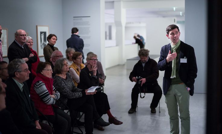 Zirkeltraining in der Berlinischen Galerie, Foto: © Harry Schnitger Foto: Viele Menschen stehen und sitzen im Ausstellungsraum und schauen auf eine Person, die spricht und auf ein Gemälde deutet.