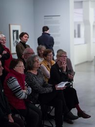 Foto: Viele Menschen stehen und sitzen im Ausstellungsraum und schauen auf eine Person, die spricht und auf ein Gemälde deutet.