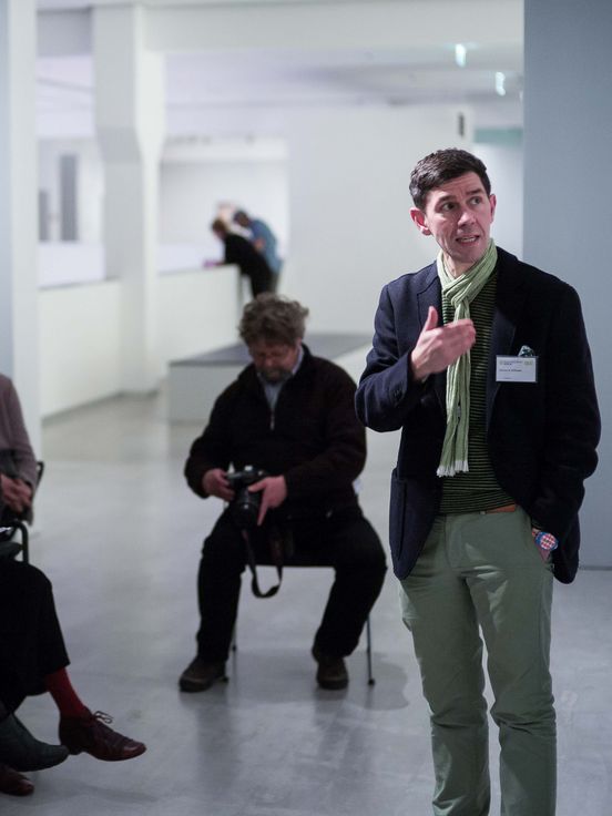 Foto: Viele Menschen stehen und sitzen im Ausstellungsraum und schauen auf eine Person, die spricht und auf ein Gemälde deutet.