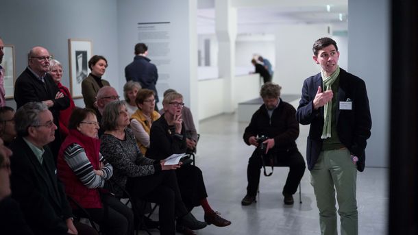 Foto: Viele Menschen stehen und sitzen im Ausstellungsraum und schauen auf eine Person, die spricht und auf ein Gemälde deutet.