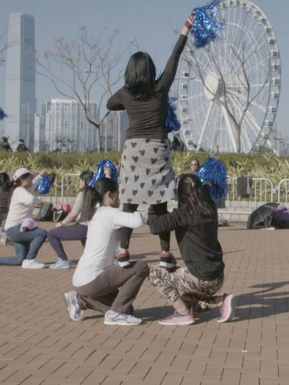 Video still: Gruppe von Menschen im Freien mit blauen Pompons, einige kniend und andere stehend, vor einem Riesenrad und Hochhäusern in einer Stadtlandschaft.