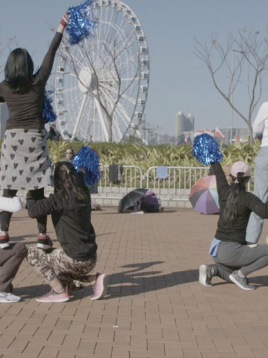 Video still: Gruppe von Menschen im Freien mit blauen Pompons, einige kniend und andere stehend, vor einem Riesenrad und Hochhäusern in einer Stadtlandschaft.
