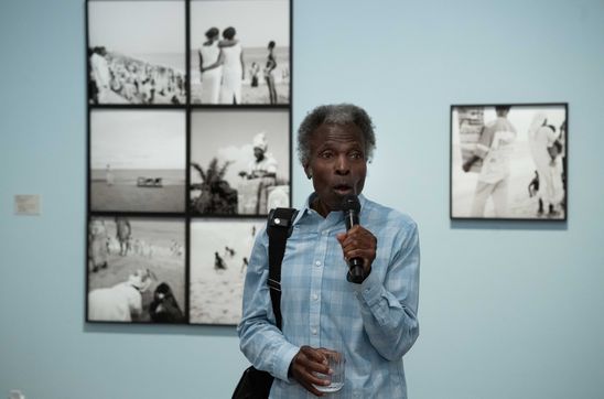 Foto: Akinbode Akinbiyi steht im Ausstellungsraum vor Schwarz-Weiß-Fotografien an einer pastell-blauen Wand und hält ein Mikrofon in der Hand.