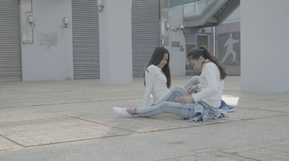 Film still: Two people in light-coloured clothing sit facing each other on the ground between two buildings.