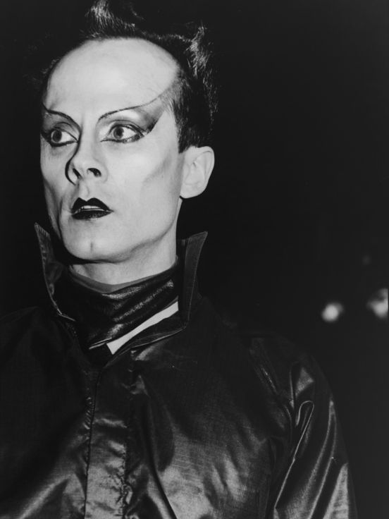 Black-and-white photograph: a portrait of Klaus Nomi wearing futuristic eye make-up, black lipstick and a leather shirt with a turned-up collar.