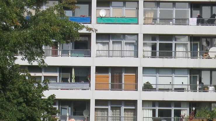 Fotografie: Hausfassade mit vielen Fenstern und Balkonen mit unterschiedlichen Vorhängen und Mobiliar. Links und unten ist der Ausblick von Baumspitzen eingerahmt..