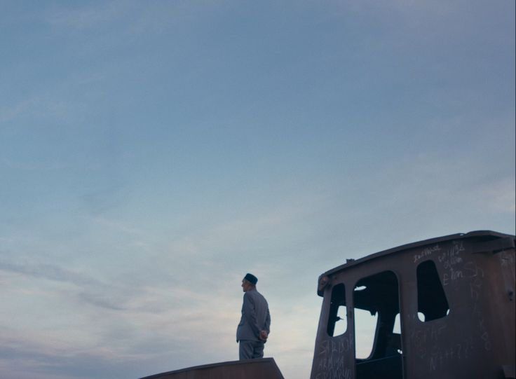 Filmstill: Seitlicher Blick auf eine ältere Person, die mit hinter dem Rücken verschränkten Armen vor dem Wrack eines Bootes steht und in die Ferne blickt.