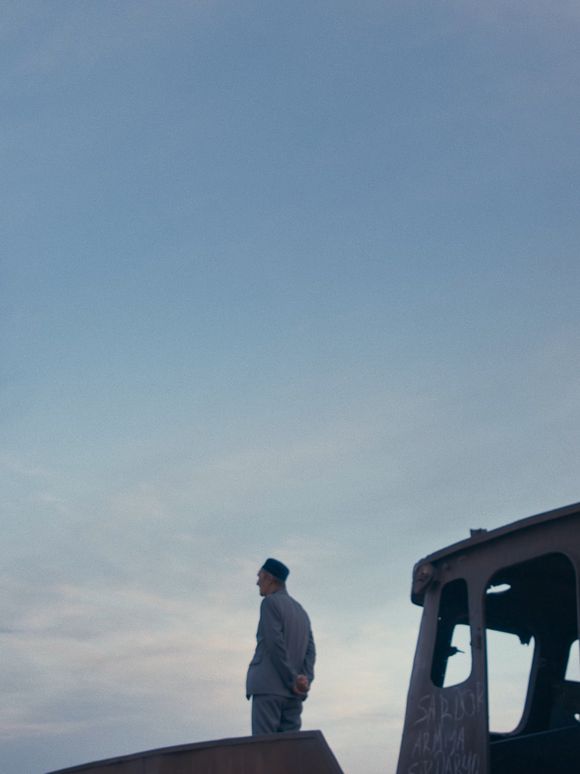 Film still: A side view of an elderly person standing in front of the wreckage of a boat, arms folded behind their back, gazing into the distance.