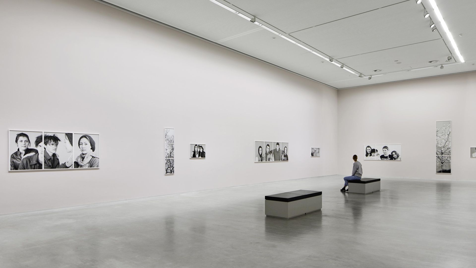 Foto: Hoher rechteckiger Ausstellungsraum mit Kunstwerken an den Wänden. Auf einer Bank sitzt eine Person.