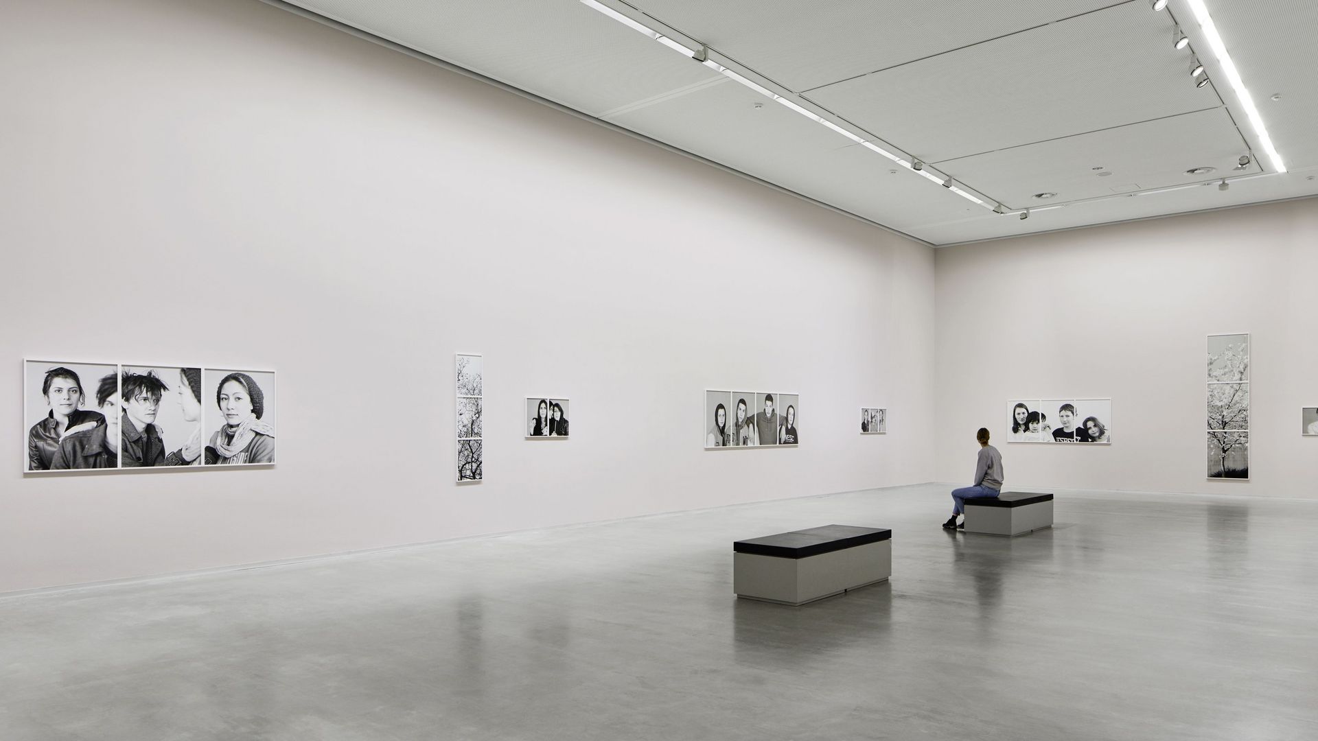 Photo: High rectangular exhibition space with artworks on the walls. A person is sitting on a bench.