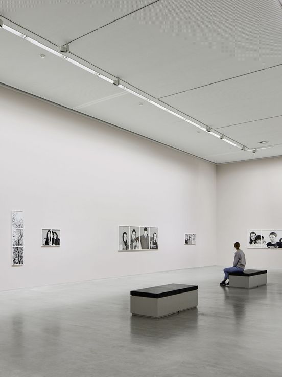 Photo: High rectangular exhibition space with artworks on the walls. A person is sitting on a bench.