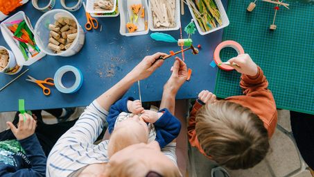 Foto: Erwachsene Person und zwei Kinder basteln gemeinsam an einem Tisch.