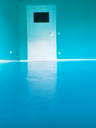 Filmstill: Blick in ein Zimmer mit leuchtend aquamarin-farbenem Boden und Wänden sowie einer weißen Tür mit dunklem Glasfenster.