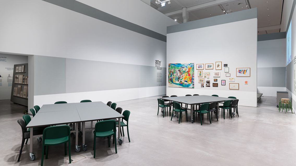 Foto: Ein großer Raum mit zwei großen Gruppentischen und vielen Stühlen. An einer freistehenden Wand hängen viele gemalte Bilder und Zeichnungen. 