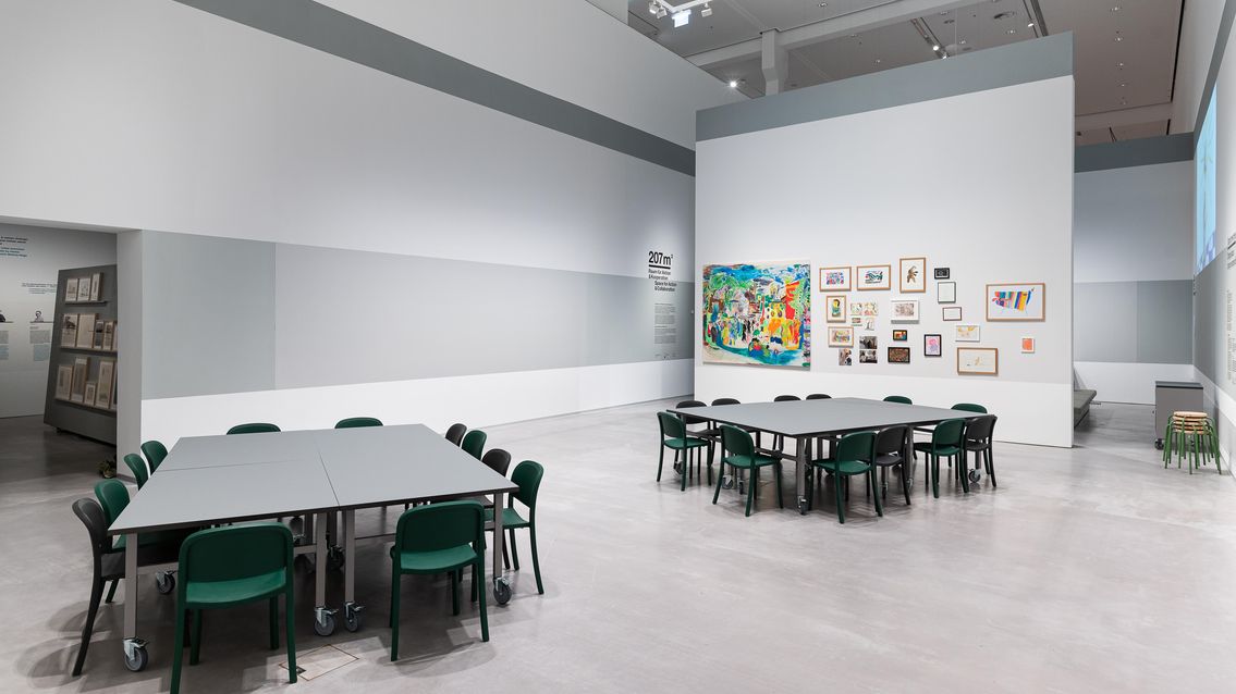 Photo: A large room with two big group tables and many chairs. On a free-standing wall hang many painted pictures and drawings.