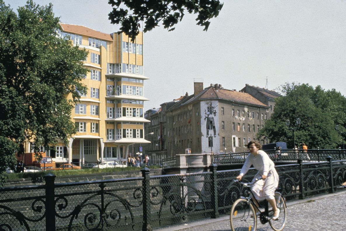 Hinrich und Inken Baller, Wohnhaus am Fraenkelufer, 1979-1984, Blick von der Uferpromenade Fotografie eines Wohnhauses an einem Flussufer. Im Vordergrund eine Radfahrerin.