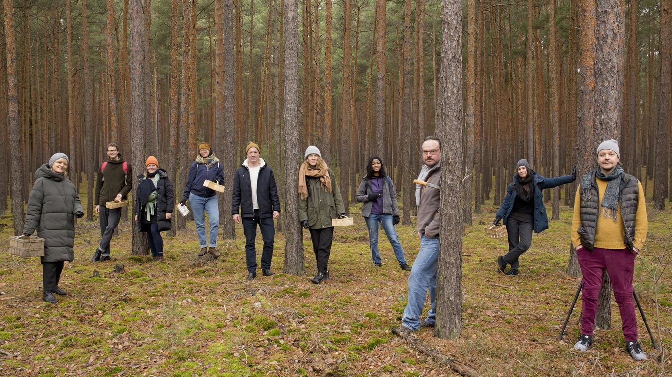 Foto: Eine Gruppe von Personen steht in einem Wald und blickt frontal in die Kamera.