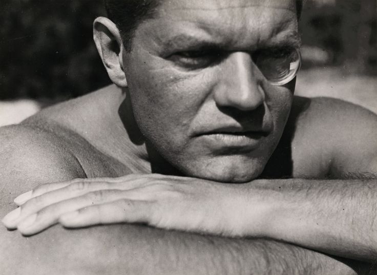 Schwarz-Weiß-Fotografie von Raoul Hausmann, der mit runder Sehhilfe im linken Auge, den Kopf auf den verschränkten Armen aufgelegt, an den Betrachtenden vorbei in die Ferne blickt.