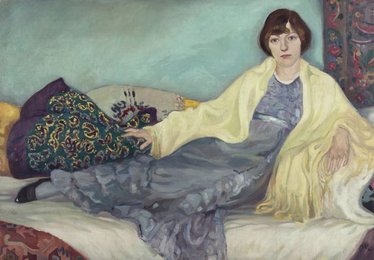 Jeanne Mammen, Die Schwester im Atelier, um 1913 Gemälde einer Frau, die auf einem Sofa sitzt. Sie trägt ein fließendes blaues Kleid und ein gelbes Tuch über den Schultern. Der Hintergrund ist türkisfarben, und um sie herum liegen gemusterte Kissen und Teppiche.